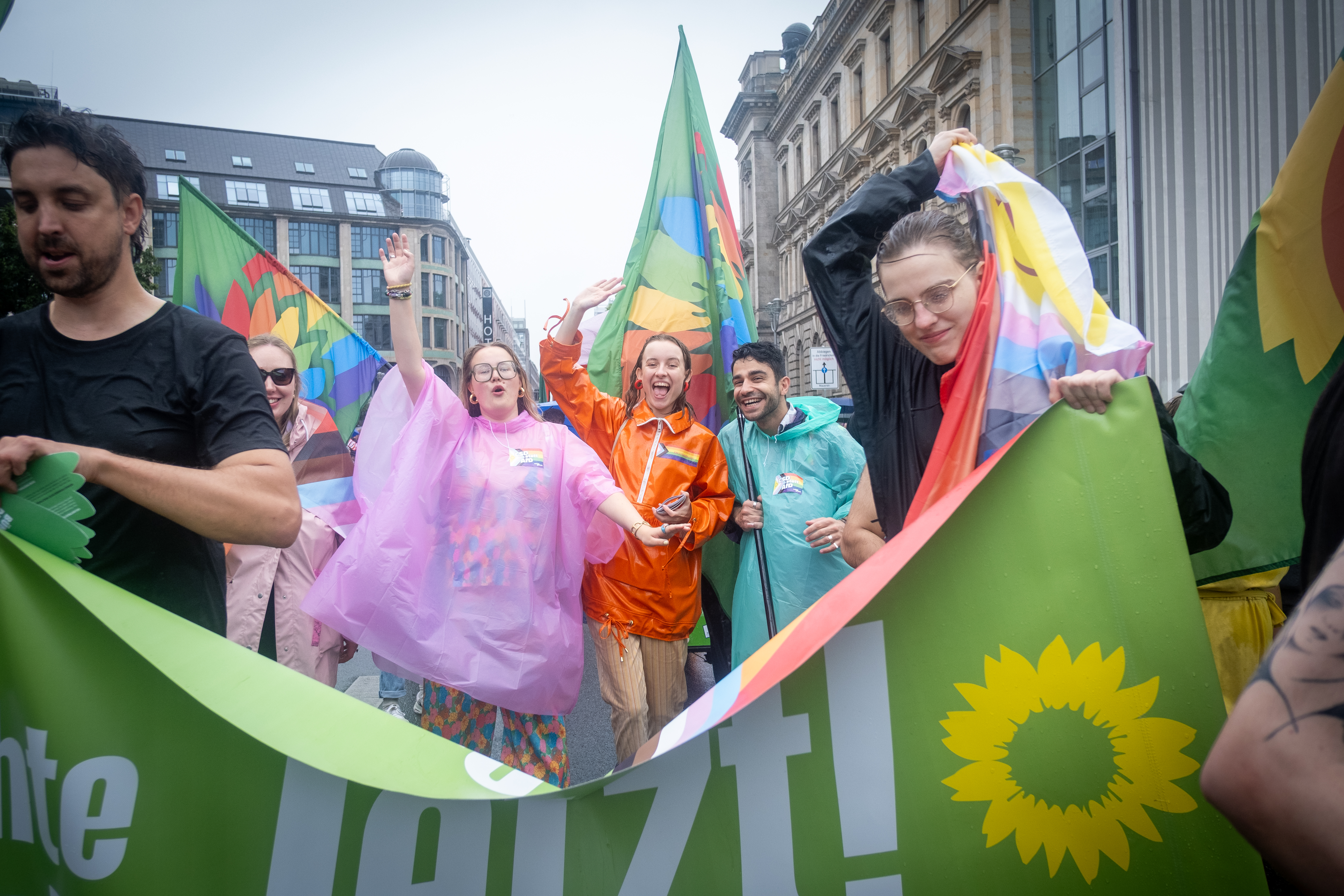 Buntgekleidete Teilnehmer*innen auf dem CSD in Berlin, sie jubeln und tragen ein grünes Banner. 