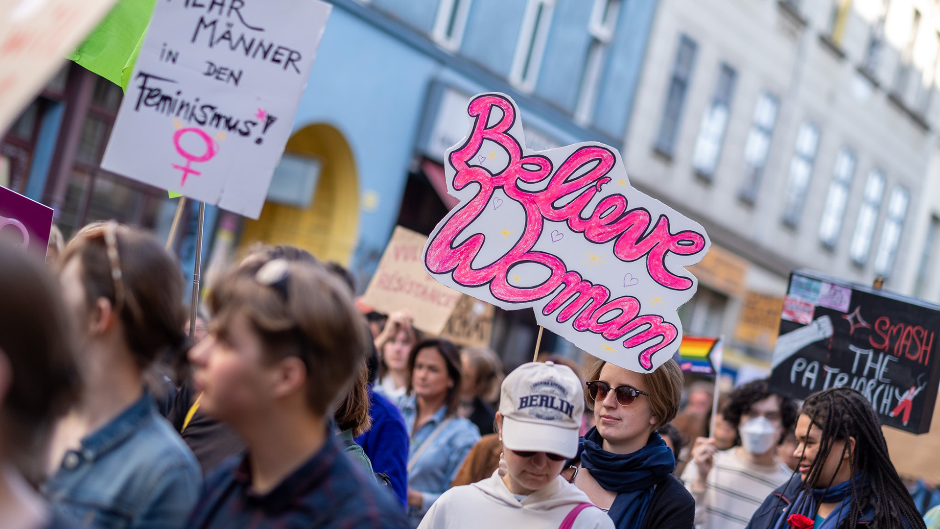 Eine Menschengruppe marschiert auf einer Demo zum 8. März. Man sieht Demoschilder mit Slogans wie "Believe Women", "Smash the Patriarchy" und "Mehr Männer in den Feminismus" 