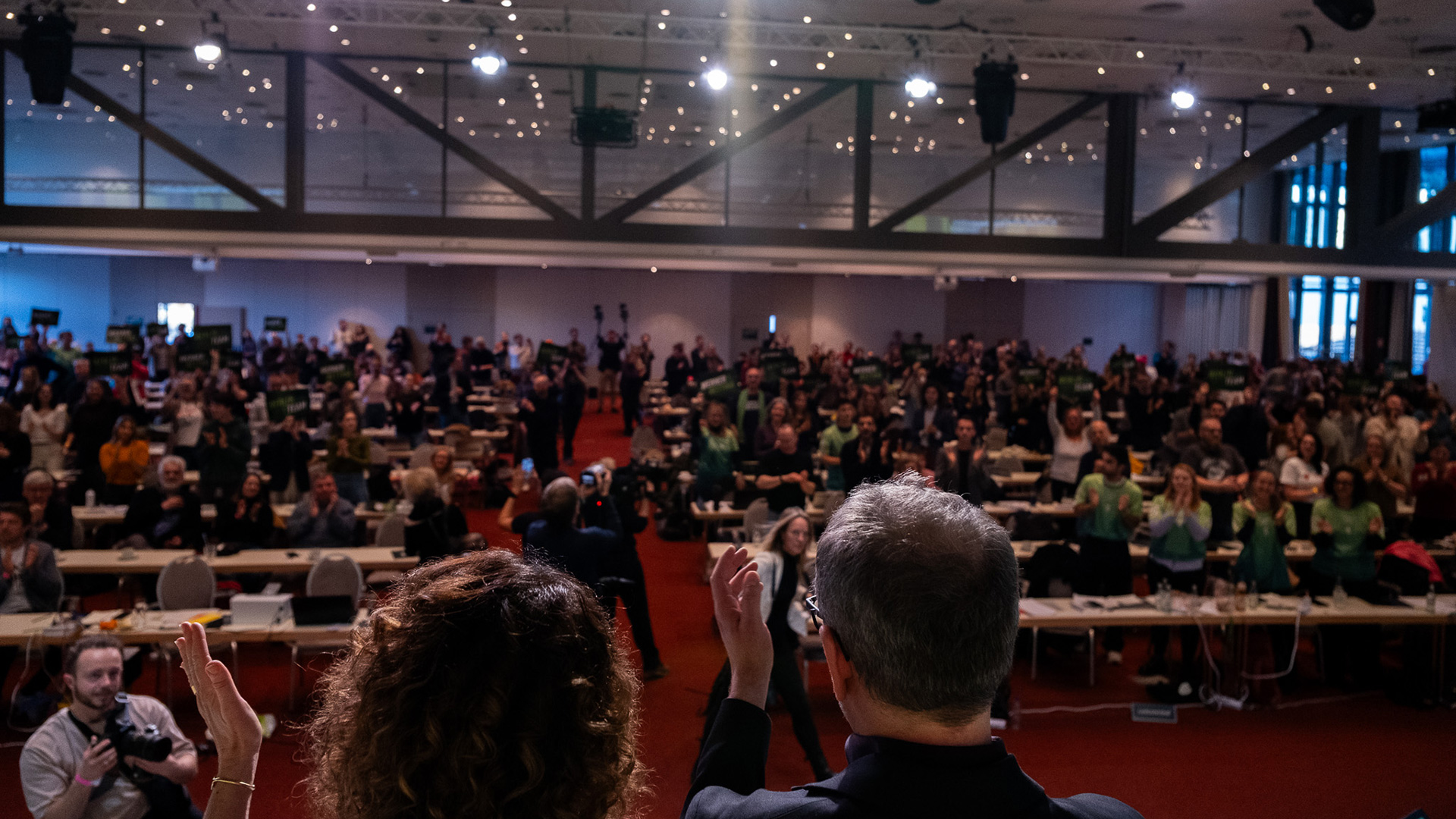 Ein voller Saal mit vielen Teilnehmer*innen, sie stehen und klatschen, die Perspektive ist von der Bühne aufgenommen, hier stehen Bettina Jarasch und Werner Graf. 