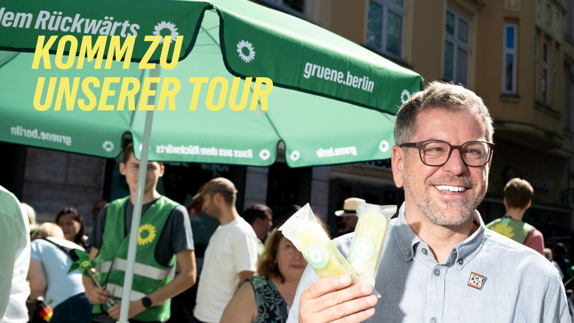 Werner Graf steht vor einem Grünen Infostand, er hält zwei Eis am Stiel in der Hand und über ihm die Schrift: Komm zu unserer Tour