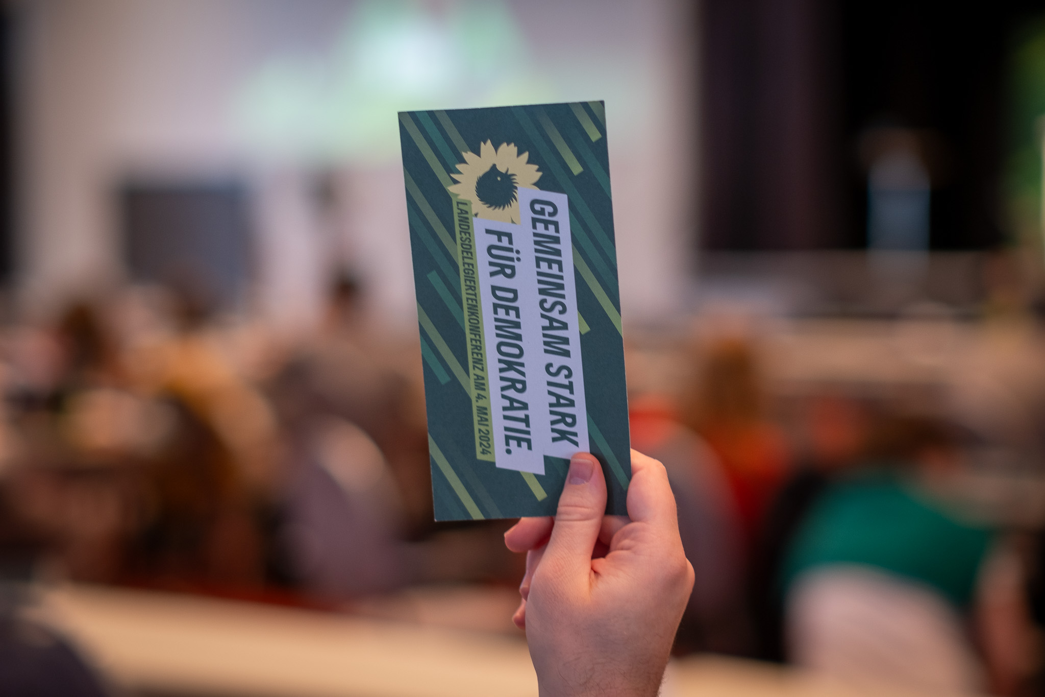 Eine Stimmkarte auf einem Grünen Parteitag in Nahaufnahme, gehalten in einer Hand. Im Hintergrund verschwommen die Teilnehmenden des Parteitags. Foto: Bündnis 90/Die Grünen Berlin