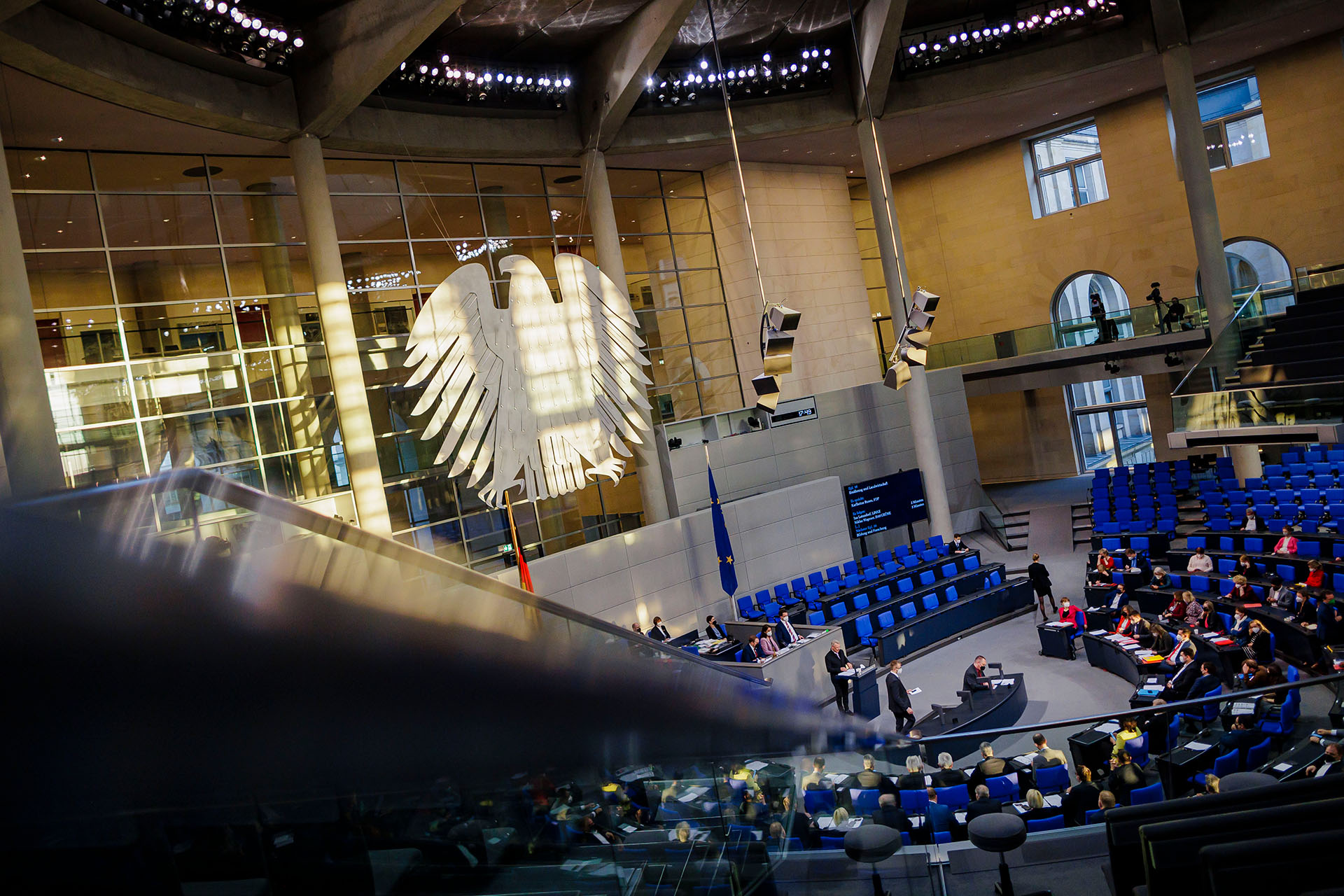 Ein Blick von schräg oben in den Plenarsaal des Deutschen Bundestages, der Bundesadler ist zu sehen sowie blaue Stuhlreihen und Personen darauf.