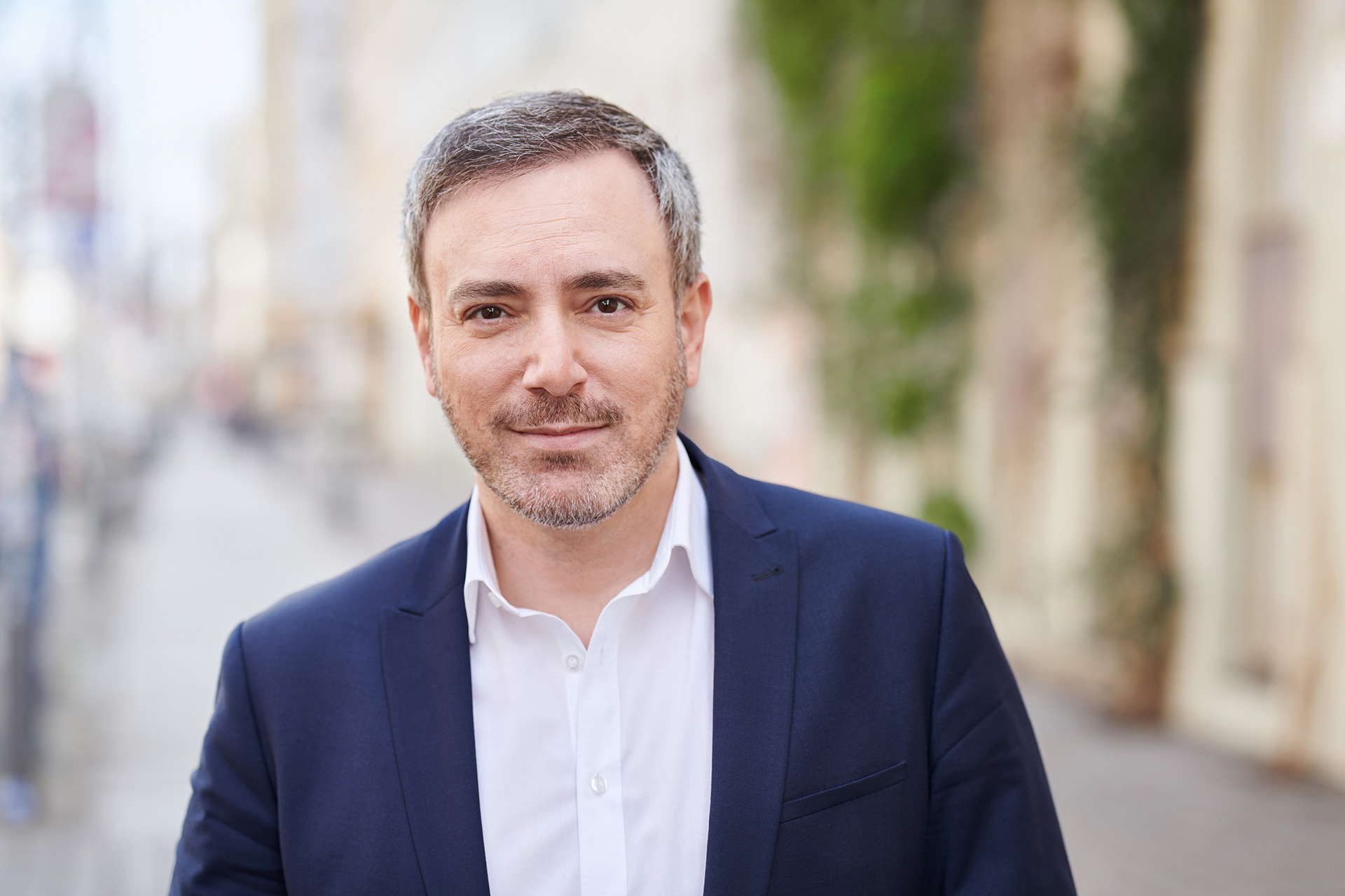 Ein Portraitfoto des grünen Abgeordneten des Europäischen Parlaments Sergey Lagodinsky. Er trägt ein weißes Hemd unter einem dunklen Sakko und lächelt in die Kamera. Er steht draußen auf einer Straße.  Foto: Jens Oellermann