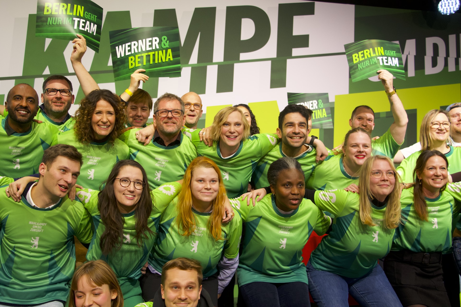 Eine große Gruppe an Menschen steht auf einer Bühne, sie sind wie ein Fußballteam aufgestellt und tragen die gleichen Trikots in Grüntönen. 