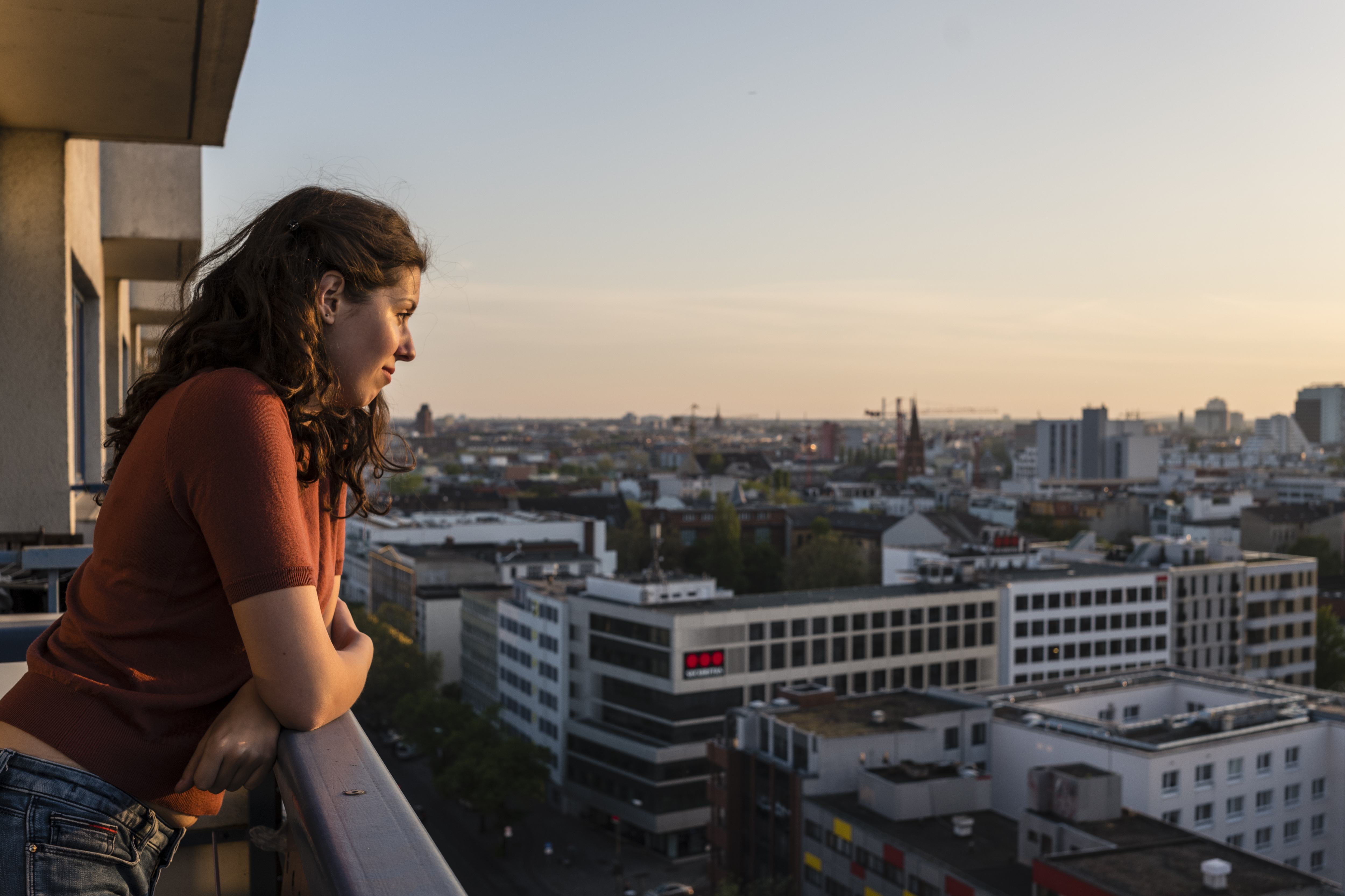 Annette Birkenfeld via Getty Images Eine junge Frau steht auf einem Balkon, lehnt sich an die Brüstung und blickt auf ein Stadtpanorama, dass den Großteil des Bildes einnimmt. Das Foto wurde bei Dämmerung aufgenommen, klar erkennbar sind Wohngebäude und -blöcke, auf die die Frau blickt. Annette Birkenfeld via Getty Images