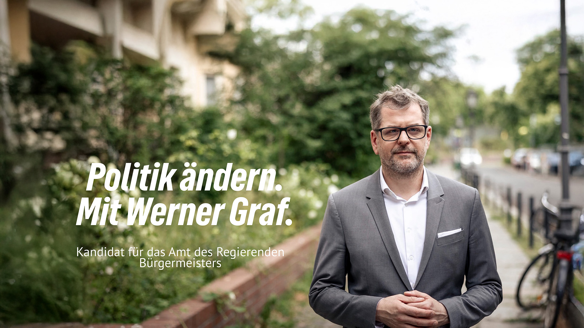 Werner Graf steht auf einer Straße, im Hintergrund Bäume, neben ihm erscheint Schrift: Politik ändern. Mit Werner Graf. Kandidat für das Amt des Regierenden Bürgermeisters. 