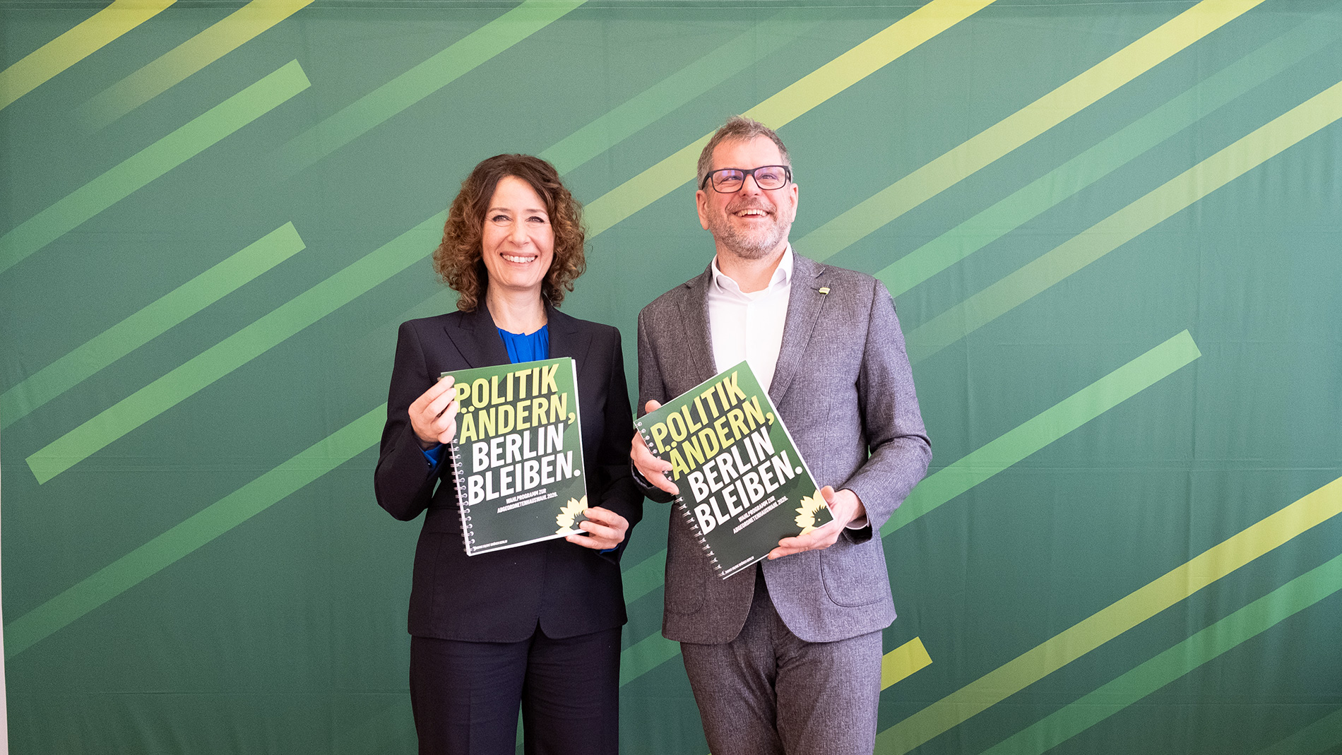 Bettina Jarasch und Werner Graf stehen mit dem Wahlprogramm in der Hand vor einer Pressewand. Auf dem Programm liest man die Aufschrift: "Politik ändern, Berlin bleiben" 