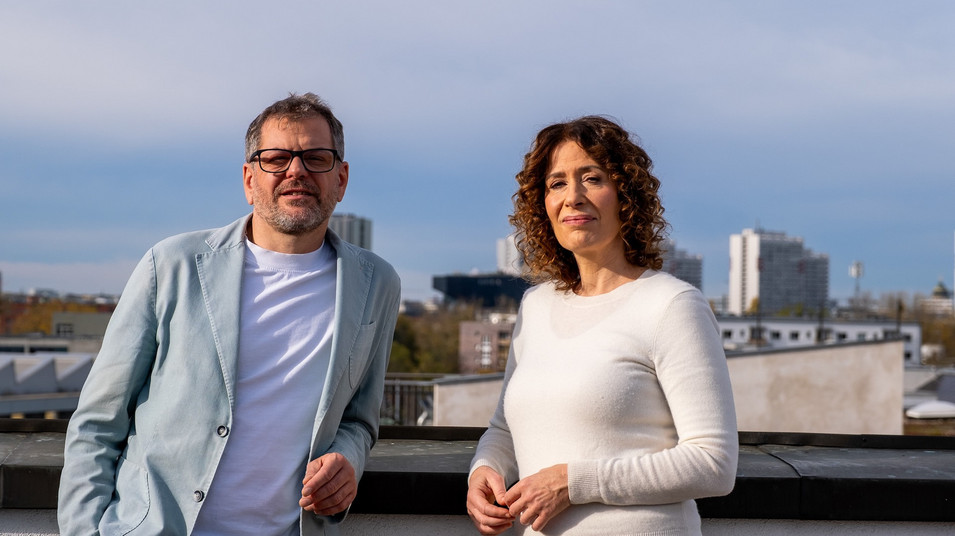 Werner Graf und Bettina Jarasch stehen auf einer Dachterrasse, hinter ihnen blauer Himmel und die Skyline von Berlin. Sie schauen in die Kamera. 