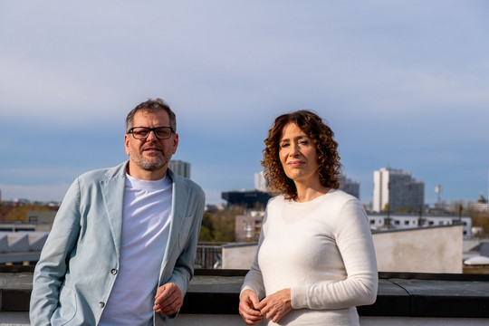 Werner Graf und Bettina Jarasch stehen auf einer Dachterrasse, hinter ihnen blauer Himmel und die Skyline von Berlin. Sie schauen in die Kamera. 