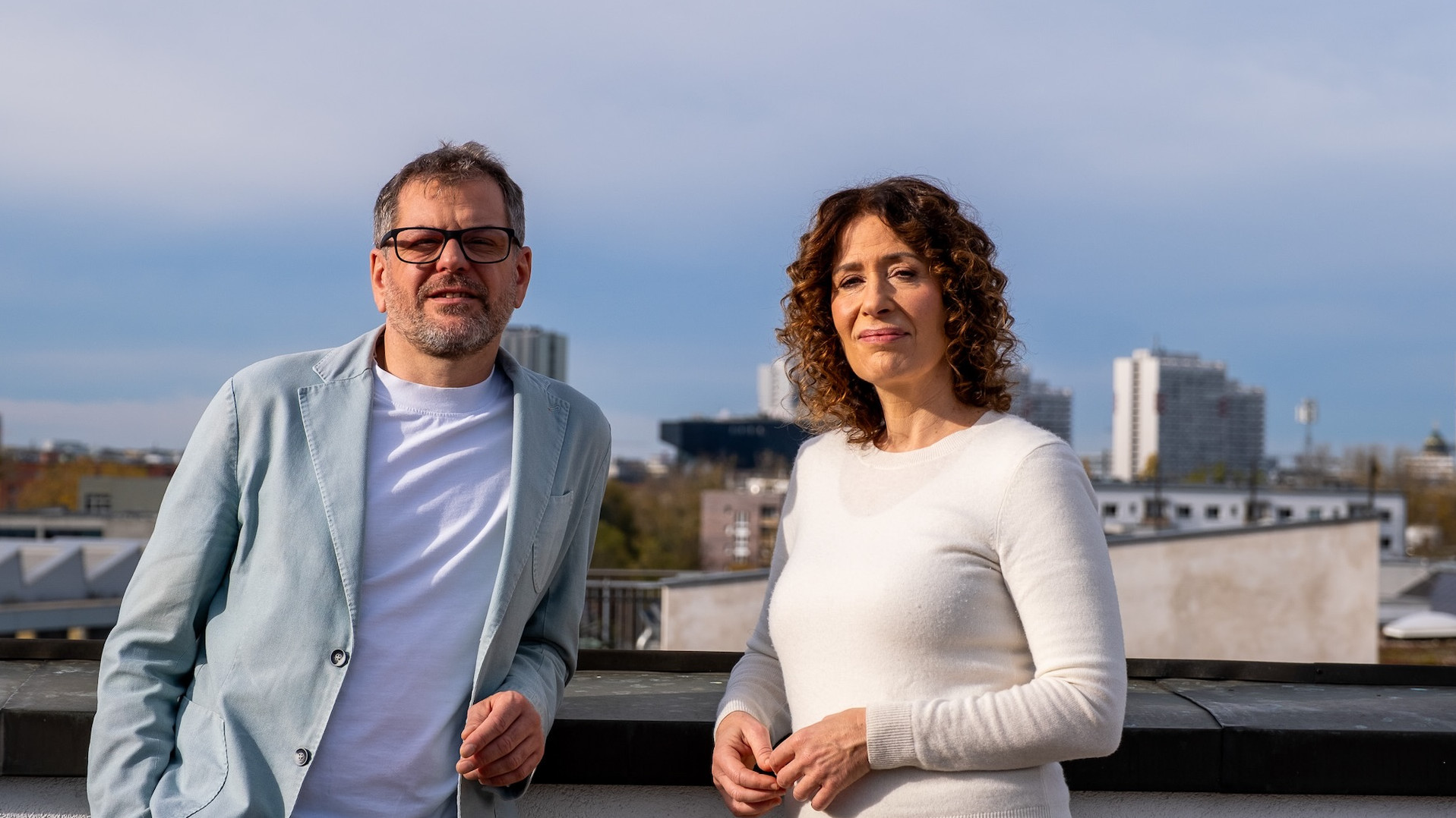 Werner Graf und Bettina Jarasch stehen auf einer Dachterrasse, hinter ihnen blauer Himmel und die Skyline von Berlin. Sie schauen in die Kamera.  