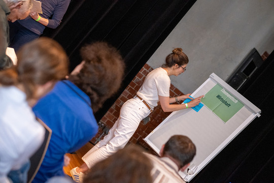 Ein gefüllter Veranstaltungsraum, eine Person steht an einem Whiteboard und notiert etwas.