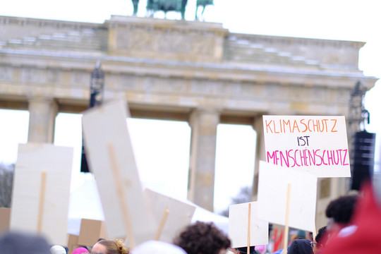Viele Personen drängen sich auf einer Demo vor dem Brandenburger Tor, man sieht Plakate mit Aufschriften wie "Klimaschutz ist Menschenschutz"