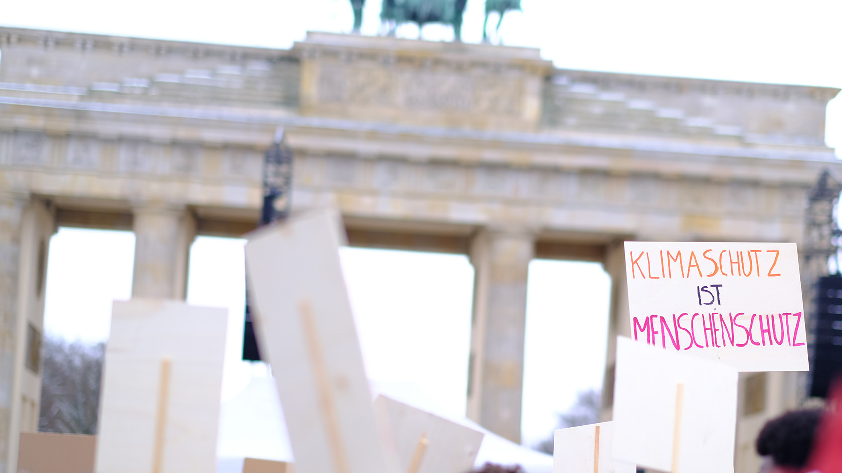 Viele Personen drängen sich auf einer Demo vor dem Brandenburger Tor, man sieht Plakate mit Aufschriften wie "Klimaschutz ist Menschenschutz" 