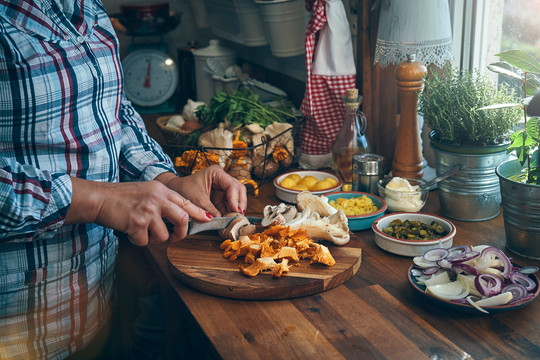 Eine Person bereitet auf einer Küchenarbeitszeile Lebensmittel zum kochen vor. Abgebildet sind die Hände und die Küche im Hintergrund. Es sind nur die Unterarme der Person erkennbar, sie schneidet auf einem Holzbrett Pilze. gmvozd via Getty Images
