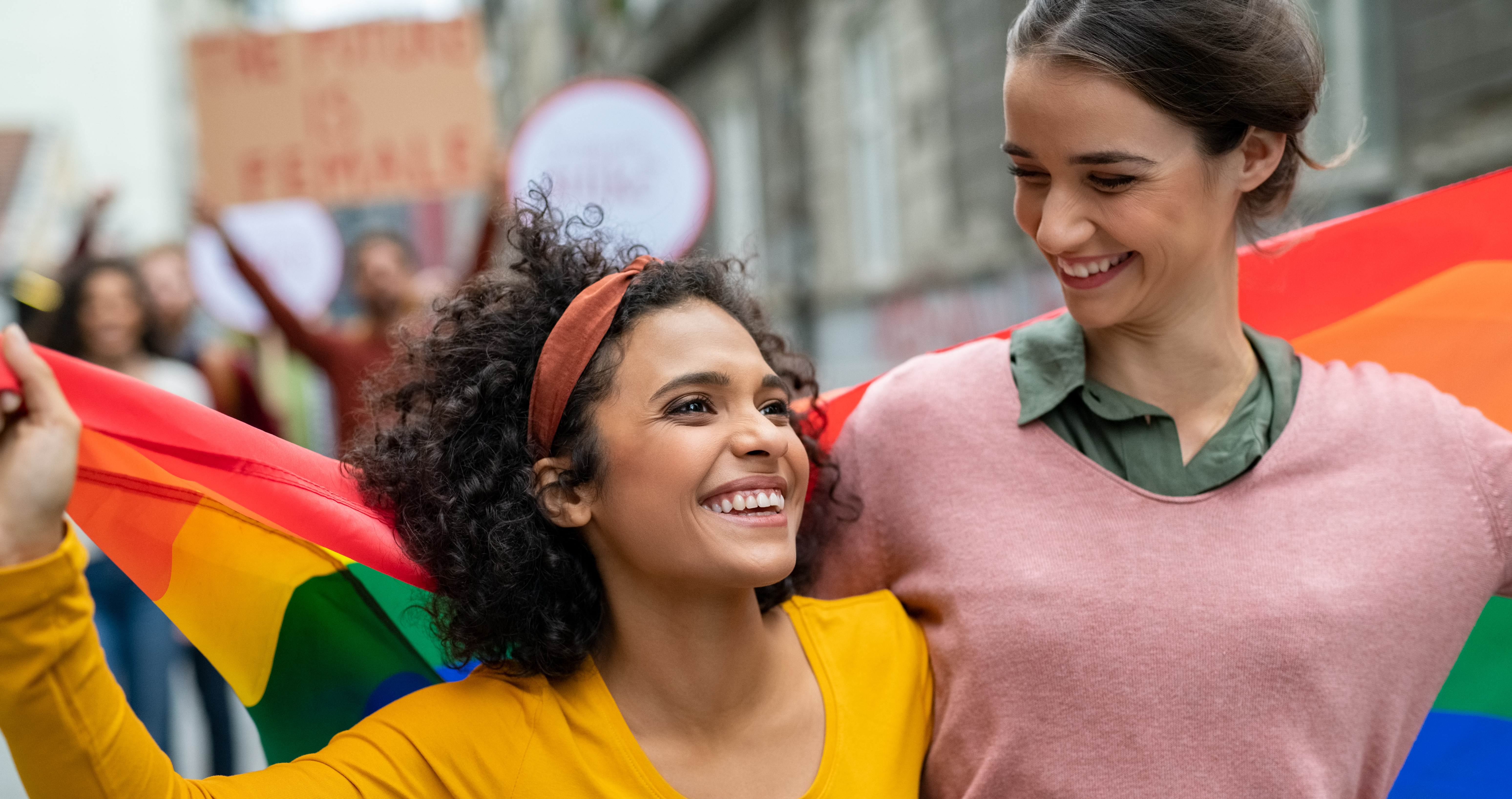 Zwei lächelnde Frauen stehen Arm in Arm an einem belebten Ort und halten eine Regenbogenfahne in den Händen. Ridofranz via Getty Images