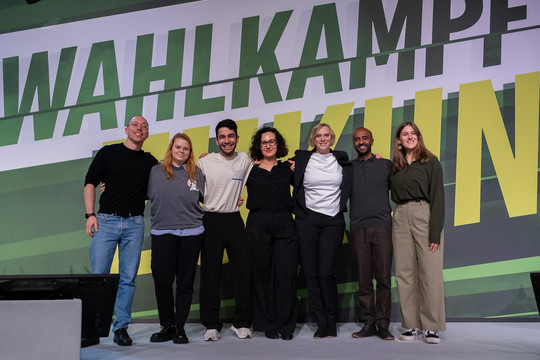 Sieben Personen stehen nebeneinander auf der Bühne: Frederic Carpenter, Landesschatzmeister, Jana Brix, Frauen- und Genderpolitische Sprecherin, Enad Altaweel, Sprecher für Vielfalt und Antidiskriminierung, Rania Al-Sahhoum, Beisitzerin, Nina Stahr, Landesvorsitzende, Philmon Ghirmai, Landesvorsitzender,  Marei Zylka, Beisitzerin. Sie lächeln in die Kamera. Hinter ihnen der Schriftzug "Wahlkampf um die Zukunft"