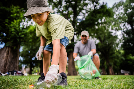 Ein Junge trägt einen Schutzhandschuh uns hebt Müll auf. Er sammelt mit seinem Vater zusammen Müll in einem Park. Der Vater ist im Hintergrund mit einem Müllbeutel zu sehen. Foto:&nbsp;Frazao Studio Latino/GettyImages