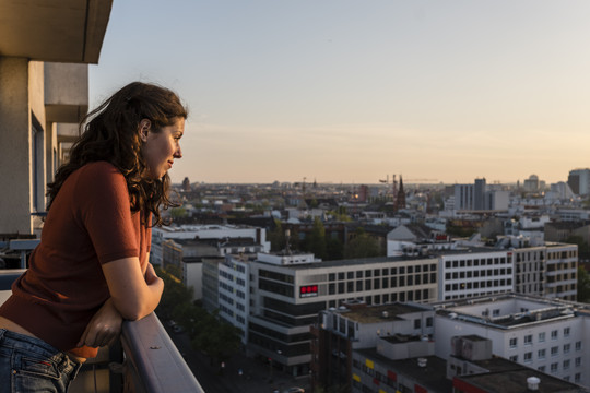 Eine junge Frau steht auf einem Balkon, lehnt sich an die Brüstung und blickt auf ein Stadtpanorama, dass den Großteil des Bildes einnimmt. Das Foto wurde bei Dämmerung aufgenommen, klar erkennbar sind Wohngebäude und -blöcke, auf die die Frau blickt. Annette Birkenfeld via Getty Images