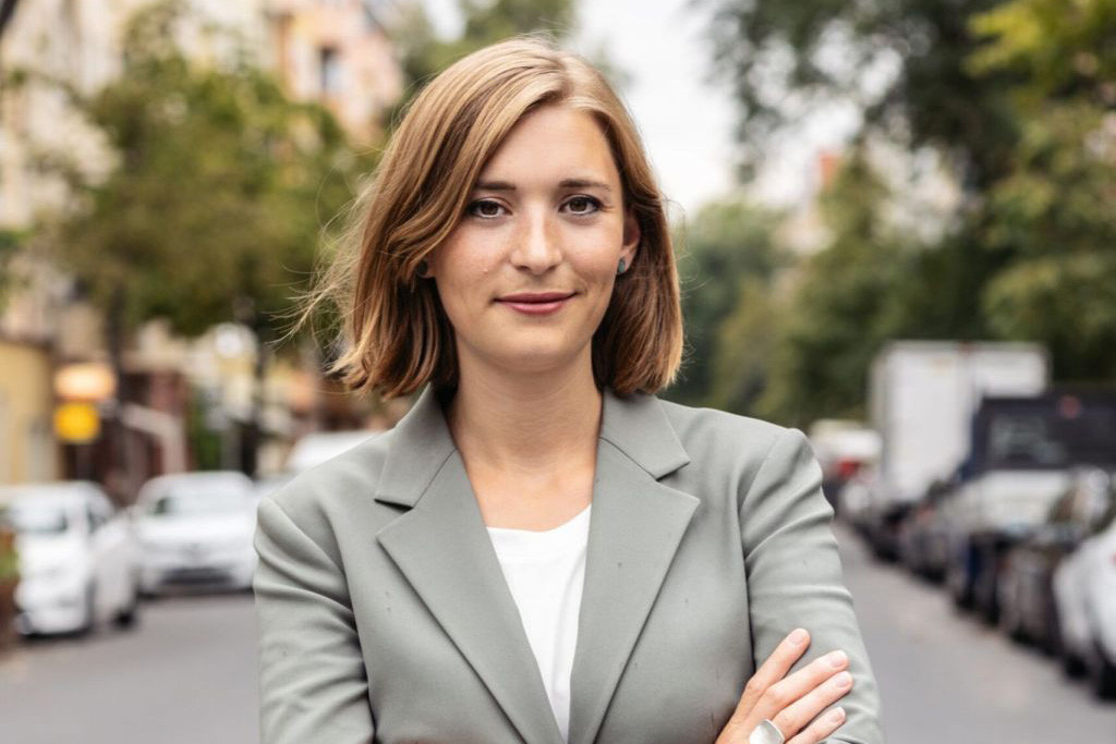 Ein Portraitfoto der Bundestagsabgeordneten Hanna Steinmüller. Sie trägt einen grauen Blazer, verschränkt ihre Arme und steht auf einer Straße.