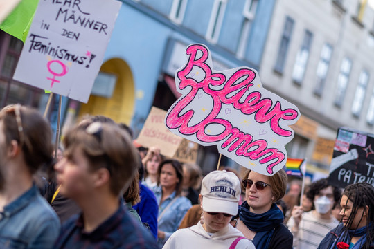 Eine Menschengruppe marschiert auf einer Demo zum 8. März. Man sieht Demoschilder mit Slogans wie "Believe Women", "Smash the Patriarchy" und "Mehr Männer in den Feminismus"