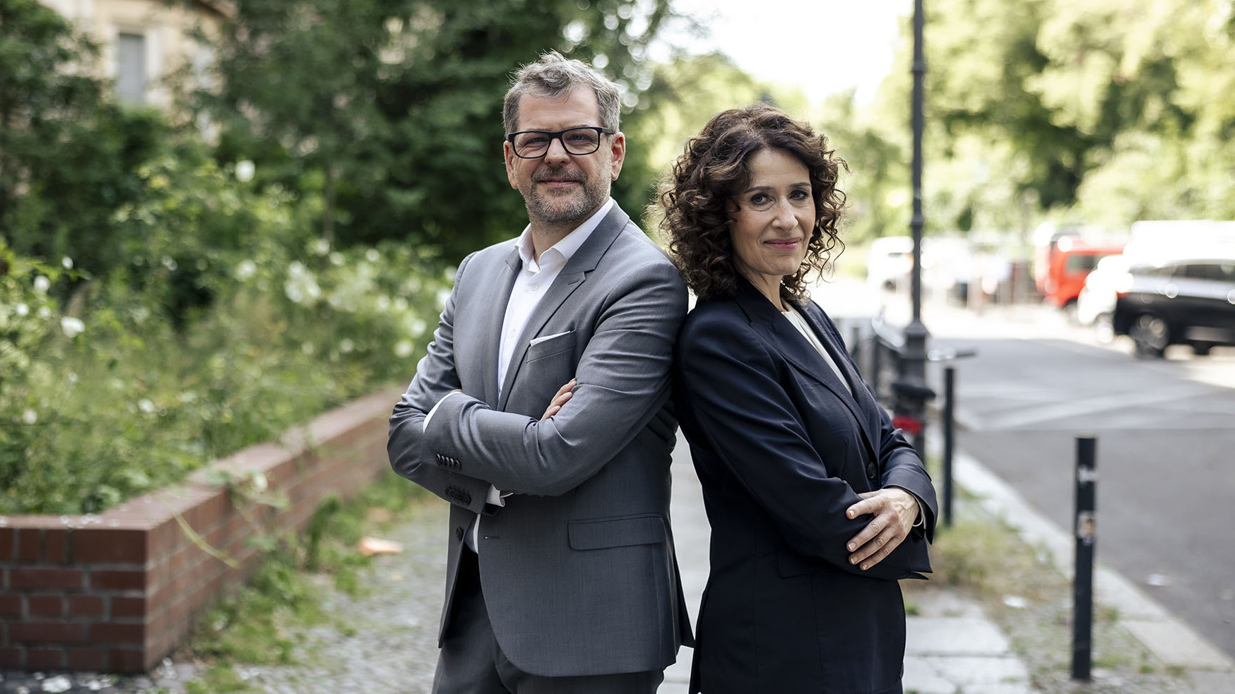 Werner Graf und Bettina Jarasch stehen Rücken an Rücken und schauen nach vorne in die Kamera. 