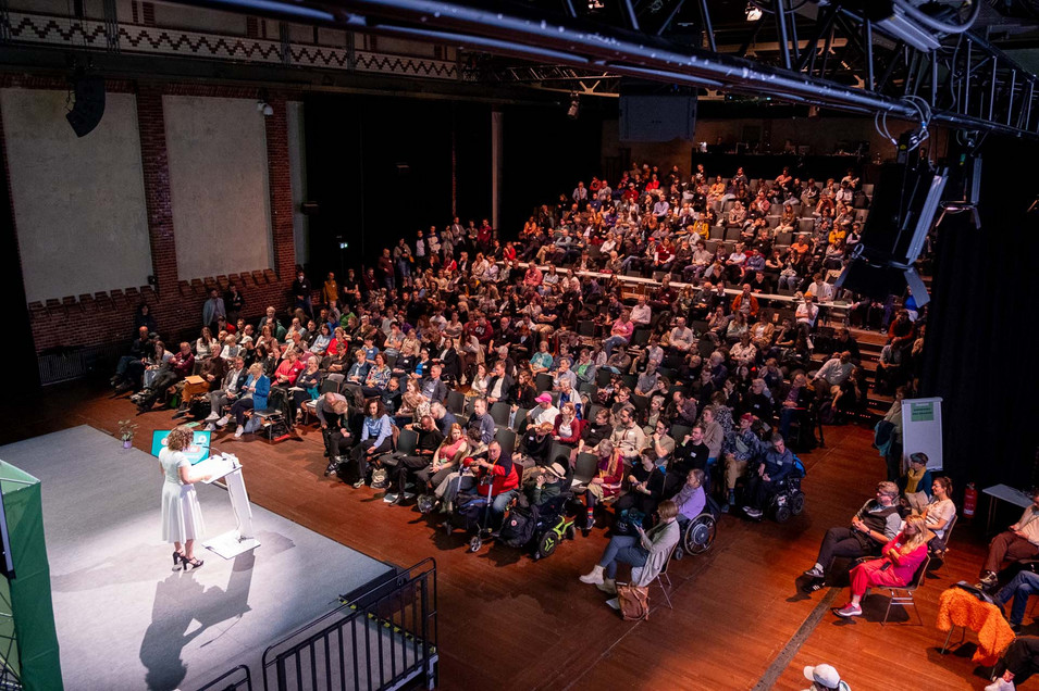 Ein Blick über einen mit Publikum gefüllten Veranstaltungssaal, vorne steht Bettina Jarrasch auf der Bühne und spricht.