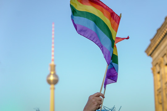 Eine Hand schwingt eine Regenbogenfahne, im Hintergrund sind blauer Himmel und der Berliner Fernsehturm zu sehen. Foto: Katrin Sauerwein/GettyImages