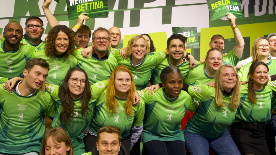 Eine große Gruppe an Menschen steht auf einer Bühne, sie sind wie ein Fußballteam aufgestellt und tragen die gleichen Trikots in Grüntönen. 