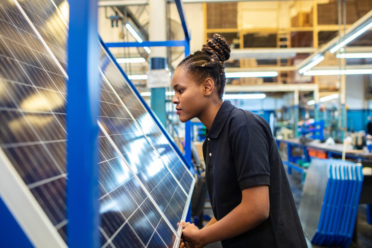 Eine junge Schwarze Frau arbeitet in einer Fertigungshalle an einem Solarpanel. Im Hintergrund sind technische Geräte und Werkzeug dargestellt. alvarez via Getty Images