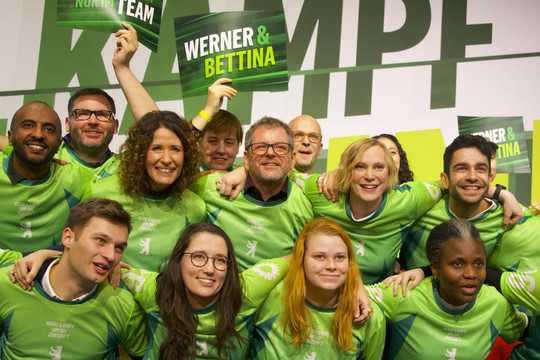 Bettina Jarasch und Werner Graf umgeben vom grünen Team Berlin auf einer Bühne, die Personen sind wie eine Fußballmannschaft aufgestellt und tragen gleiche Trikots. Foto: Bündnis 90/Die Grünen Berlin