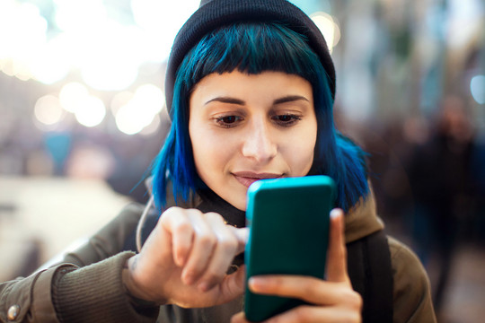 Eine junge Frau mit blauen Haaren schaut auf ihr Smartphone und tippt auf dessen Bildschirm.