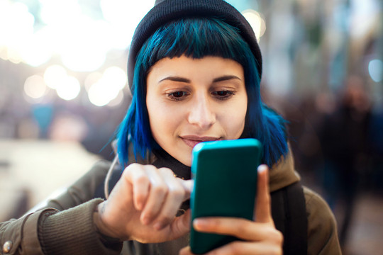 Eine junge Frau mit blauen Haaren schaut auf ihr Smartphone und tippt auf dessen Bildschirm. Marco Piunti via Getty Images