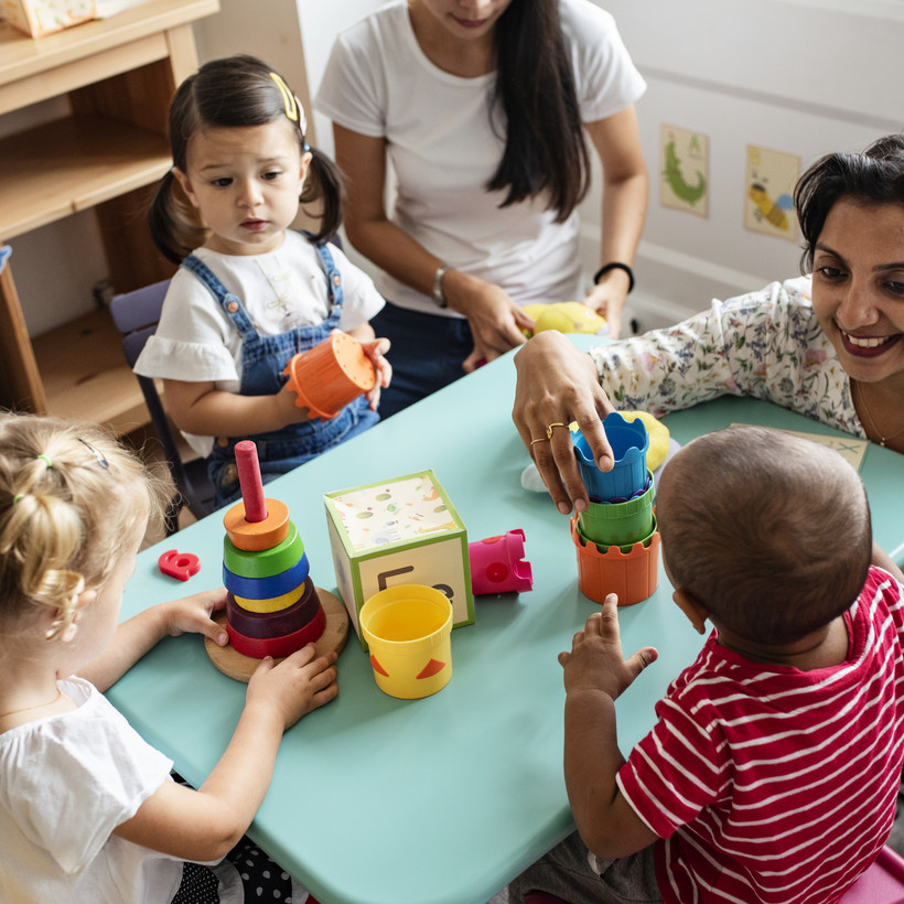Eine Erzieherin sitzt in einer Kita mit Kinder an einem Tisch, vor ihnen steht Spielzeug. Foto: Rawpixel via GettyImages