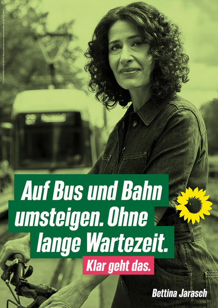 Die Bürgermeisterkandidatin Bettina Jarasch im Portrait während sie ein Fahrrad schiebt. Im Hintergrund ist eine Straßenbahn. Die Bildaufschrift lautet "Auf Bus und Bahn umsteigen. Ohne lange Wartezeit. Klar geht das.". Daneben ist das Logo von Bündnis 90/Die Grünen abgebildet. 