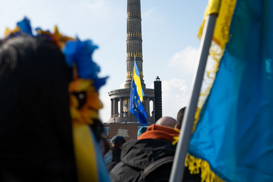 Eine Menschenmenge auf einer Demonstration an der Siegessäule in Berlin. Einzelne Personen sind nicht erkennbar. Im Vordergrund und im Hintergrund sind viele Fahnen und andere Gegenstände mit ukrainischen Flaggen oder in den Farben der Ukraine. Foto: Daniel Meißner