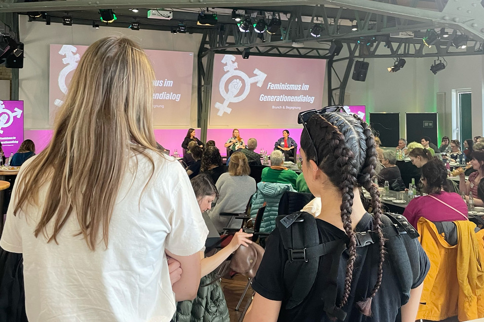 Zwei Personen stehen mit dem Rücken zur Kamera, eine Person trägt ein Baby in einer Trage. Sie schauen auf den vollen Saal und die Bühne.