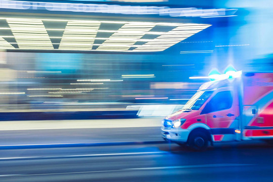Ein Rettungswagen fährt an einem erleuchteten Gebäude vorbei, der Rettungswagen ist auf dem Foto unscharf zu erkennen.