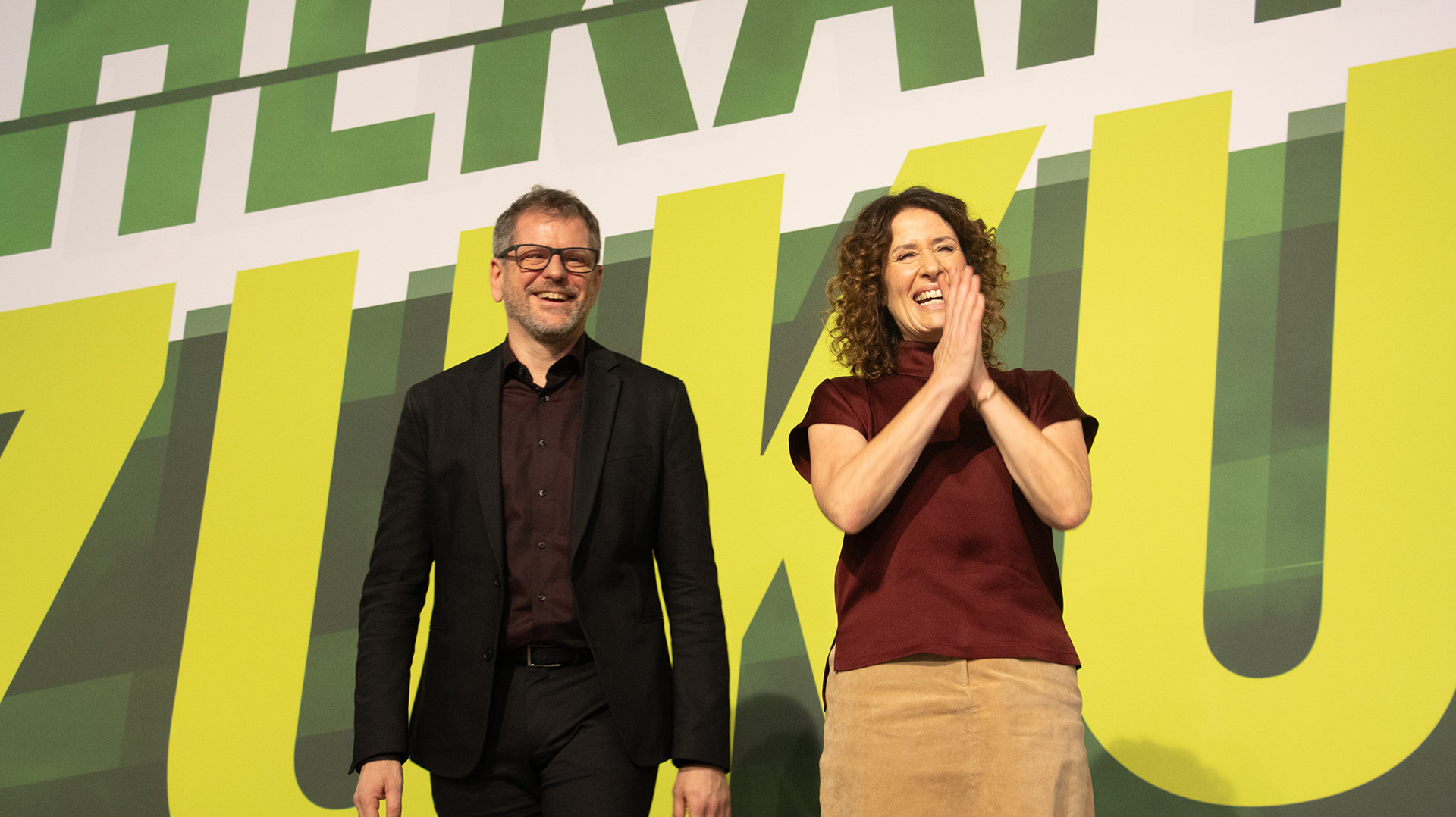 Werner Graf und Bettina Jarasch stehen nebeneinander auf einer Bühne und jubeln. Im Hintergrund sieht man einen großen Schriftzug. 